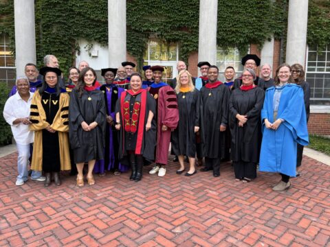 Faculty and staff in academic regalia stand in front of brick building with white columns covered in ivy