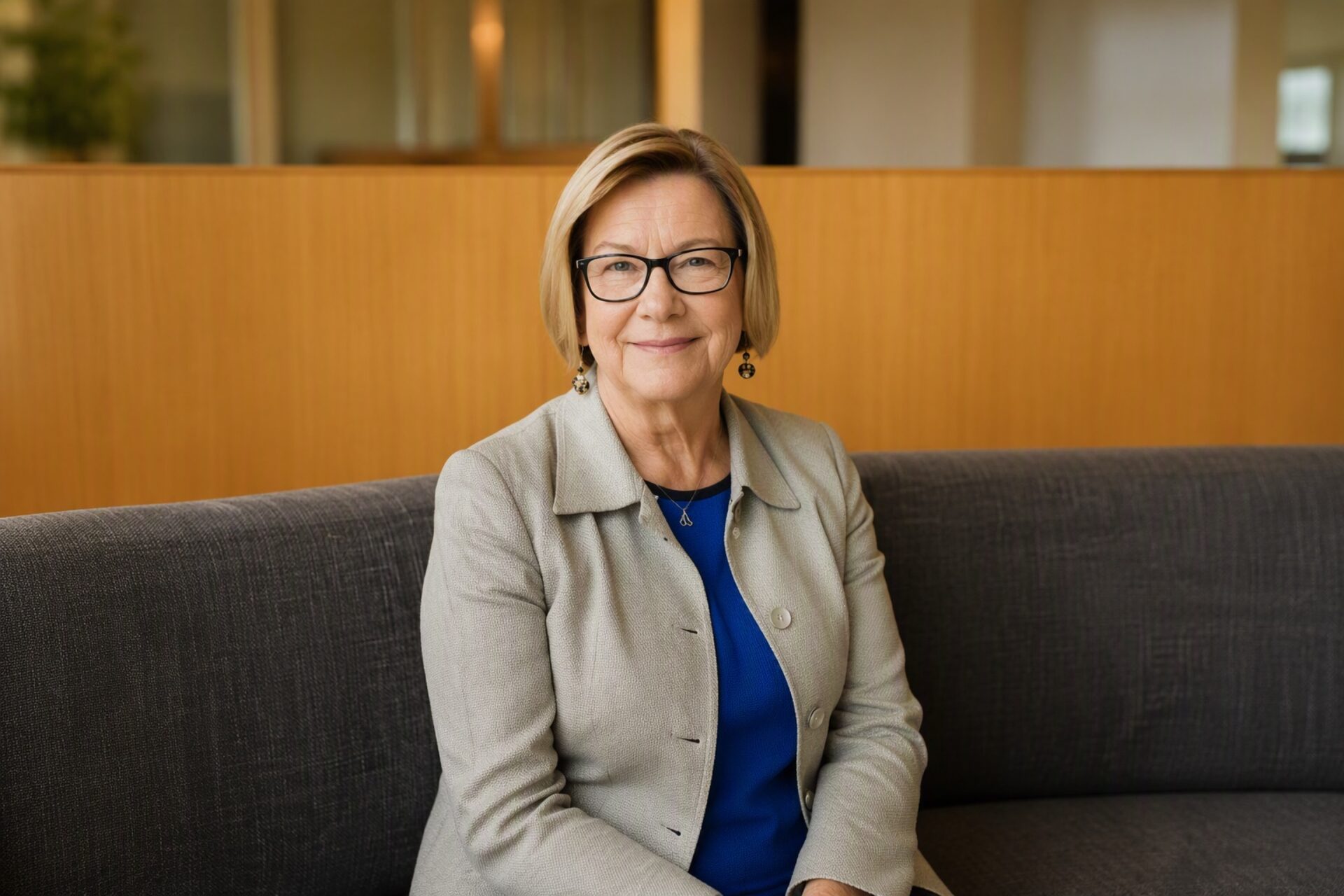 White woman with short blond hair wearing tan suit and blue blouse smiles gently at the camera in professional headshot