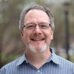 White man with goatee and glasses in blue striped button down shirt smiles at the camera