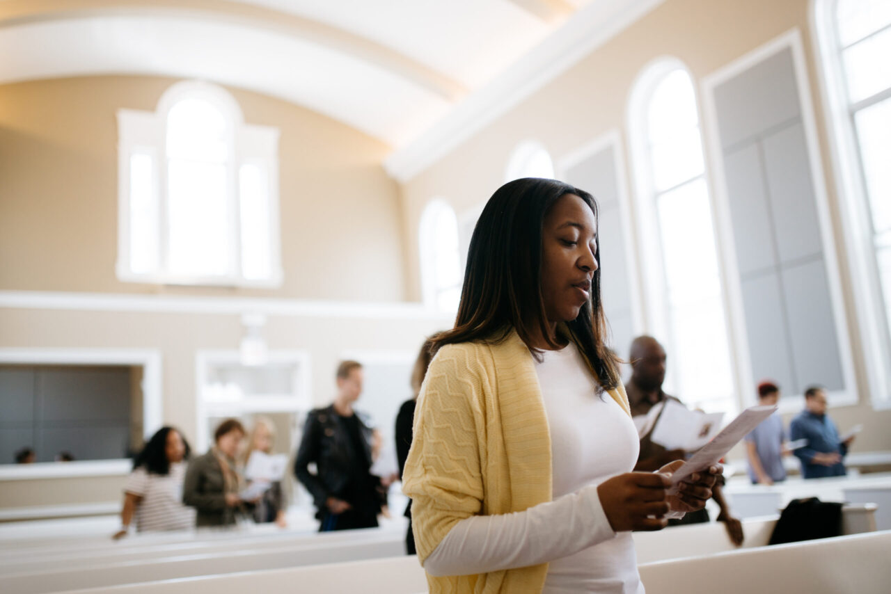 Black woman with yellow sweater and white shirt sings in a light-filled chapel