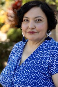 Latina woman with short dark hair wearing dangly earrings and blue and white blouse smiles at the camera