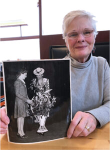Older white woman wearing glasses holds up black and white photograph
