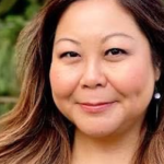 Asian woman with golden-brown hair smiles at the camera while standing in front of greenery