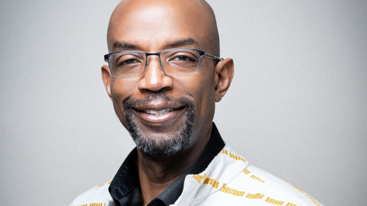 Bald Black man with goatee and glasses smiles at the camera