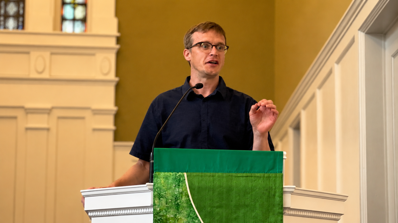 White man wearing glasses and blue polo shirt preaches from a pulpit in a chapel