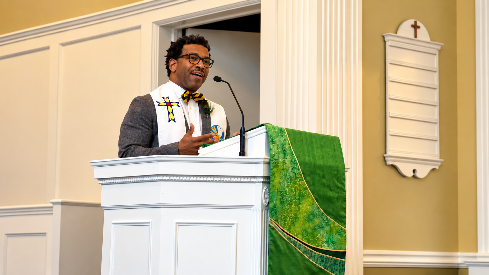 Black clergyman wearing bowtie and stole standing at a white pulpit delivering a sermon